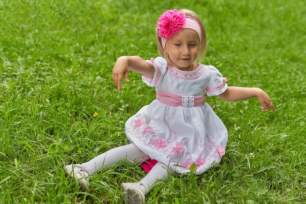 Retrato de uma menina em um vestido e um laço na cabeça. Criança sentada na grama verde com os braços abertos