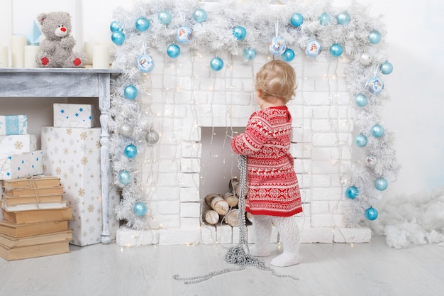 retrato de uma menina em um vestido vermelho perto de uma lareira de tijolos em casa de Natal decorada. férias de Natal dentro de casa com brinquedos e caixa de presente