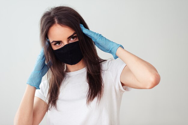 Retrato de uma menina em uma máscara médica preta e uma camiseta branca sobre um fundo branco, as mãos nas luvas perto da cabeça. sintomas de uma doença de coronovírus. ficar em casa conceito