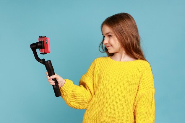 Retrato de uma menina encantadora blogueira transmitindo ao vivo usando steadicam e telefone celular vestindo suéter amarelo estilo casual Tiro de estúdio interno isolado em fundo azul