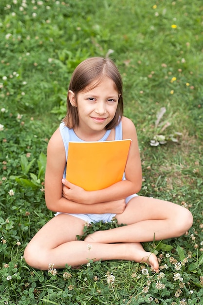 Retrato de uma menina encantadora de 8 a 10 anos de idade em uma camiseta e shorts com um livro amarelo nas mãos, em um prado verde verão, imagem filtrada. Educação