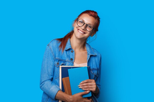 Retrato de uma menina estudante caucasiana usando óculos, sorrindo e segurando livros de exercícios isolados.