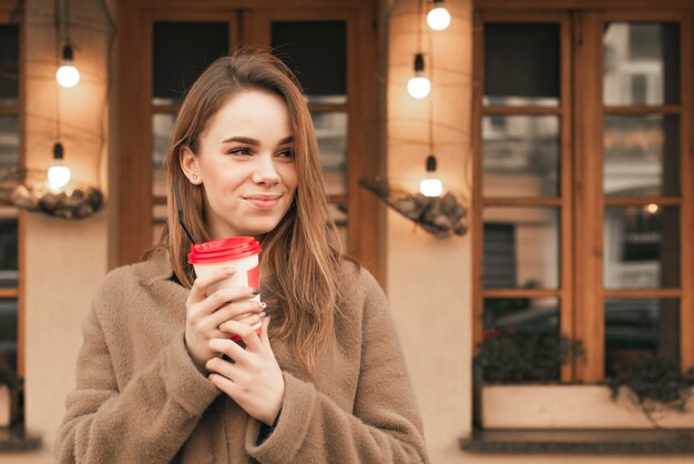 Retrato de uma menina feliz fica no fundo de uma parede marrom com uma xícara de café nas mãos, veste roupas de primavera, olha de soslaio e sorri