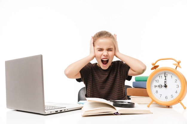 Retrato de uma menina infeliz usando um laptop prateado enquanto estuda e lê livros na aula, isolados sobre uma parede branca