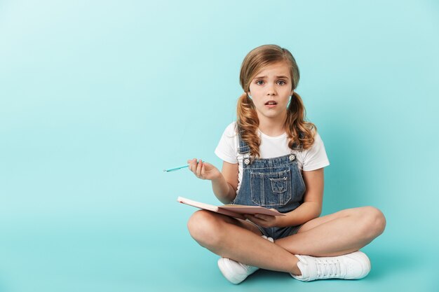 Retrato de uma menina isolada sobre uma parede azul, escrevendo em um caderno