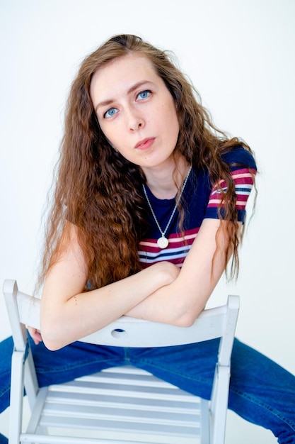 Retrato de uma menina jovem e séria sentada em uma cadeira em um fundo branco com cabelo comprido vestindo uma camiseta apertada e jeans