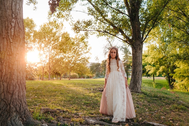 Retrato de uma menina loira com cabelo encaracolado com vestido de comunhão caminhando por entre algumas árvores