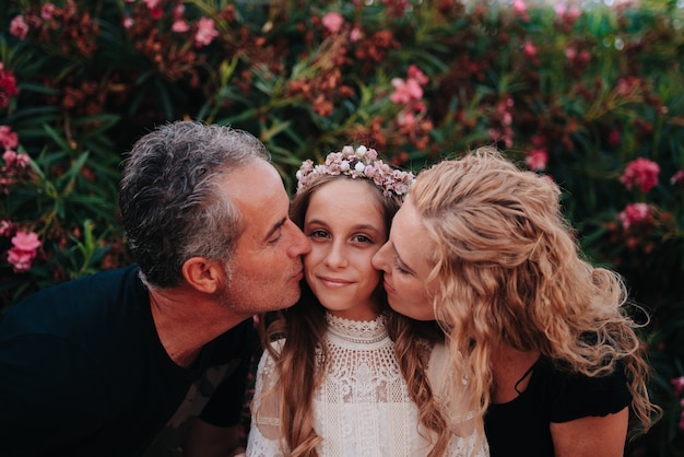 Foto retrato de uma menina loira com cabelo encaracolado vestida com um vestido de comunhão dando um beijo nos pais