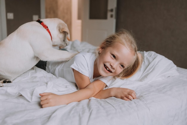 Retrato de uma menina loira deitada na cama com seu cachorro amado em seus braços. garota de camiseta branca e jeans e um animal de estimação estão deitados na roupa de cama branca. espaço para texto. foto de alta qualidade