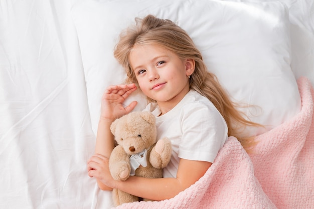 retrato de uma menina loira deitada na cama com um ursinho de pelúcia sob uma vista de cima de um cobertor rosa