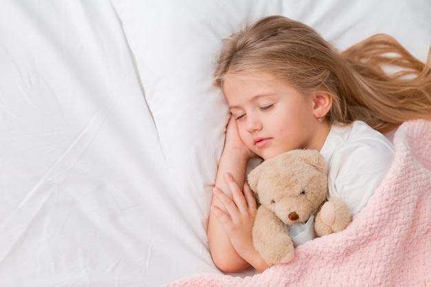 retrato de uma menina loira dormindo em uma cama com um ursinho de pelúcia sob um cobertor rosa