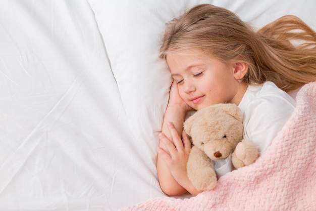 Foto retrato de uma menina loira dormindo em uma cama com um ursinho de pelúcia sob um cobertor rosa
