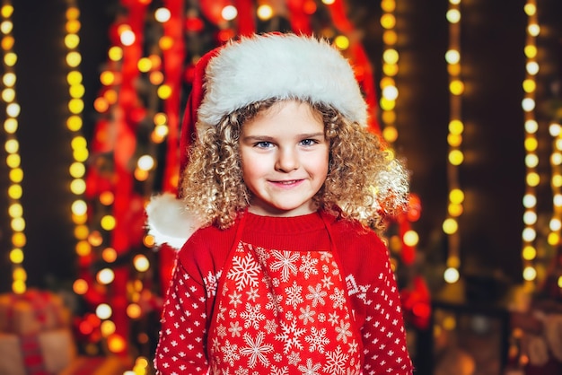 Retrato de uma menina loira encaracolada com chapéu de Papai Noel em luzes desfocadas fundo conceito de Natal