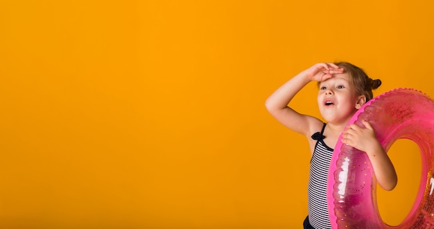 Retrato de uma menina loira feliz em um maiô listrado segurando um círculo inflável rosa em uma superfície amarela com espaço para texto