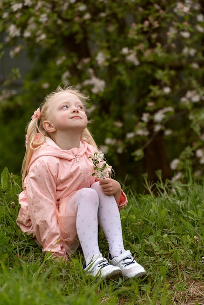 Retrato de uma menina loira sentada na grama e olhando para cima Criança em jardim em flor Quadro vertical