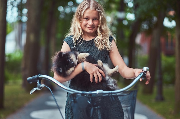 Retrato de uma menina loira sorridente em um vestido casual, segura cachorro spitz fofo. Andar de bicicleta em um parque.