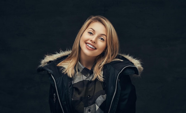 Retrato de uma menina loira sorridente em uma jaqueta de inverno e camisa de lã, de pé em um estúdio. isolado em um fundo escuro.