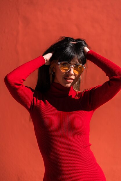 Retrato de uma menina morena sorrindo em um vestido laranja e óculos de sol na primavera, aproveitando uma manhã ensolarada. Caucasiano muito um fundo vermelho