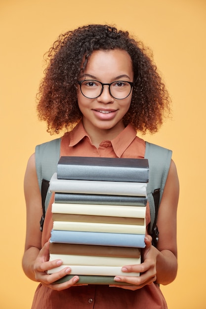 Foto retrato de uma menina negra inteligente sorridente em óculos, segurando uma pilha de livros enquanto se preparava para o seminário.