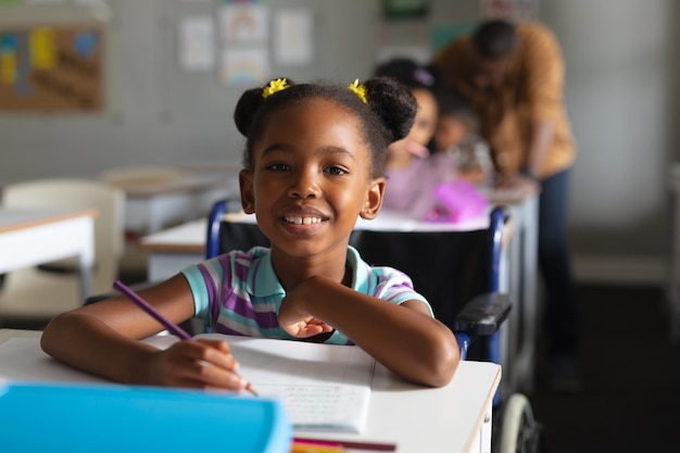 Retrato de uma menina primária afro-americana sorridente estudando sentada em cadeira de rodas na secretária
