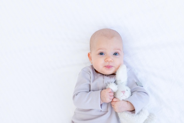 Retrato de uma menina recém-nascida em um terno de algodão em uma cama branca em casa deitada e sorrindo com uma lebre de pelúcia na mão, um lugar para texto