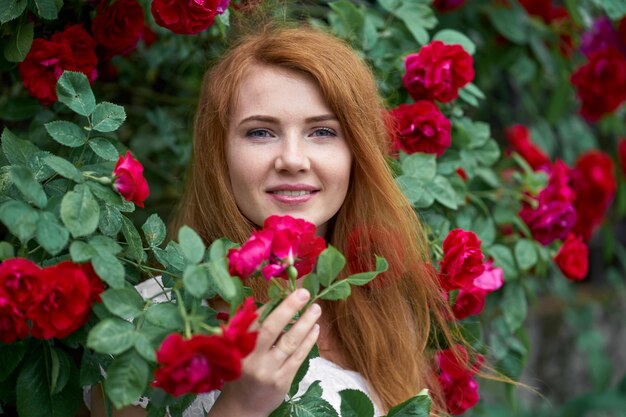 Retrato de uma menina ruiva bonita vestida com um vestido de luz branca sobre um fundo de rosas florescendo.