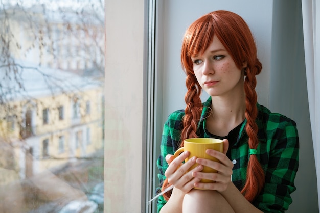 Retrato de uma menina ruiva em uma camisa verde, sentada no parapeito da janela e segurando uma caneca de chá amarela.