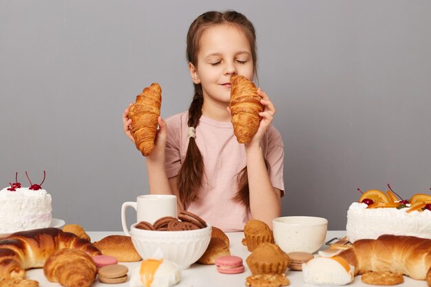 Retrato de uma menina satisfeita com tranças sentada à mesa isolada sobre fundo cinza, cheirando delicioso croissant com os olhos fechados, desfrutando de um doce jantar