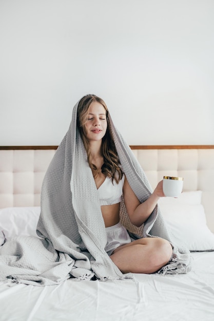 Foto retrato de uma menina sentada na cama com uma xícara de café coberta com um cobertor