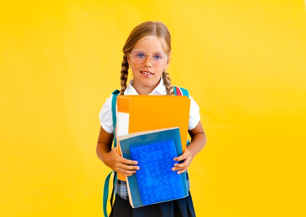 Retrato de uma menina sorridente com uma mochila segurando um caderno sobre fundo amarelo