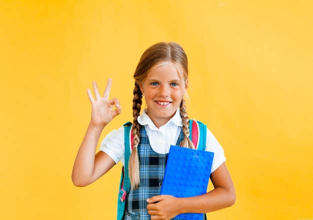 Retrato de uma menina sorridente com uma mochila segurando um caderno sobre fundo amarelo