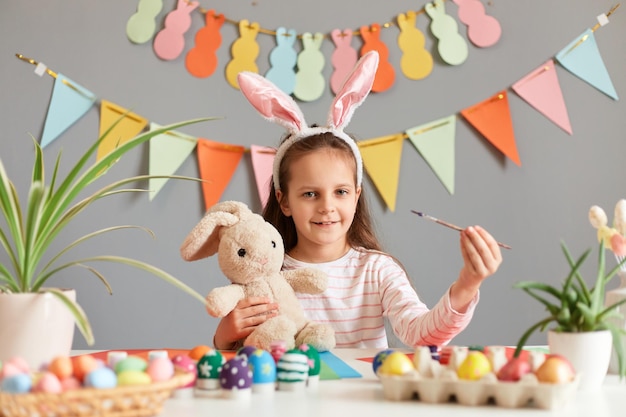 Retrato de uma menina sorridente e alegre vestida com orelhas de coelho, sentada à mesa, segurando nas mãos o brinquedo e pintando ovos de Páscoa contra a parede cinza decorada