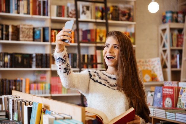 Retrato de uma menina sorridente e feliz tirando uma selfie enquanto está sentado na biblioteca