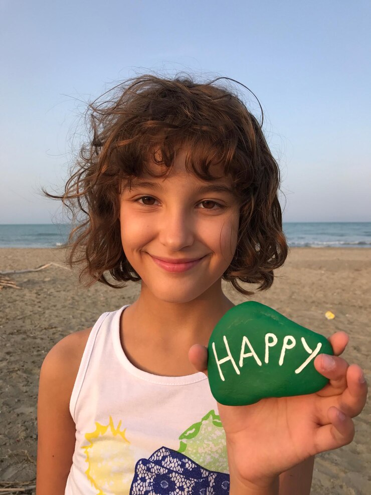 Foto retrato de uma menina sorridente mostrando texto em uma rocha enquanto está de pé na praia contra o céu