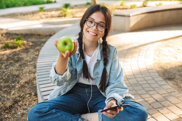 Retrato de uma menina sorridente positivo bonito jovem estudante usando óculos, sentado no banco ao ar livre no parque natural, usando telefone celular conversando, ouvindo música com fones de ouvido, segurando a maçã.