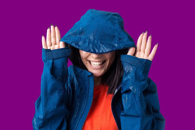 Foto retrato de uma menina sorridente vestida com capa de chuva azul em gotas posando com capuz em fundo cinza em um estúdio