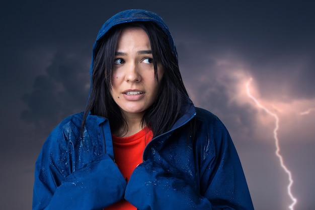 Retrato de uma menina sorridente, vestida com capa de chuva azul em gotas posando com capuz no contexto de um céu sombrio com relâmpagos