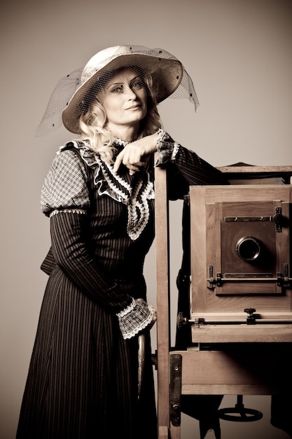 Retrato de uma misteriosa jovem positiva com um vestido longo modesto e chapéu se passando perto de uma velha câmera de daguerreótipo. estilização do conceito de foto vintage retrô.