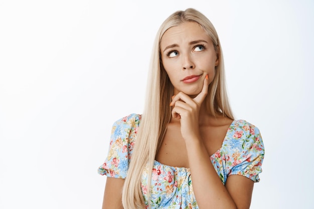 Retrato de uma modelo feminina loira pensativa olhando para cima e pensando em pé de vestido sobre fundo branco copiar espaço