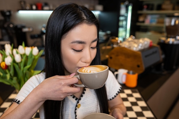 Retrato de uma morena calma bebendo cappuccino com espuma de um copo de cerâmica no café