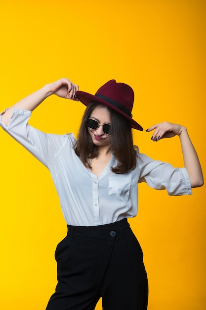 Retrato de uma morena com um chapéu de camisa branca e óculos em um fundo amarelo