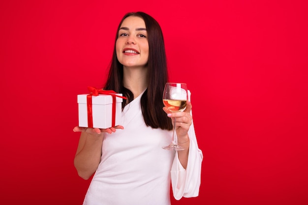Retrato de uma morena de vestido branco com um copo de vinho e uma caixa de presente em um fundo vermelho Uma jovem celebra o Ano Novo