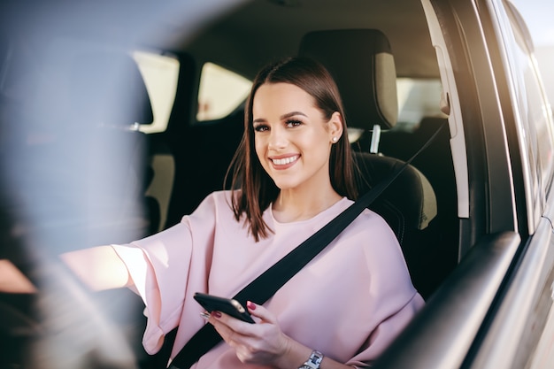 Retrato de uma morena linda com um grande sorriso, dirigindo o carro, usando telefone inteligente e olhando para a câmera.