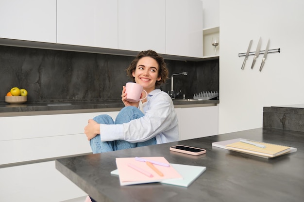 Retrato de uma morena sorridente sentada em casa bebe uma xícara de chá na cozinha relaxa aproveita o dia de folga i