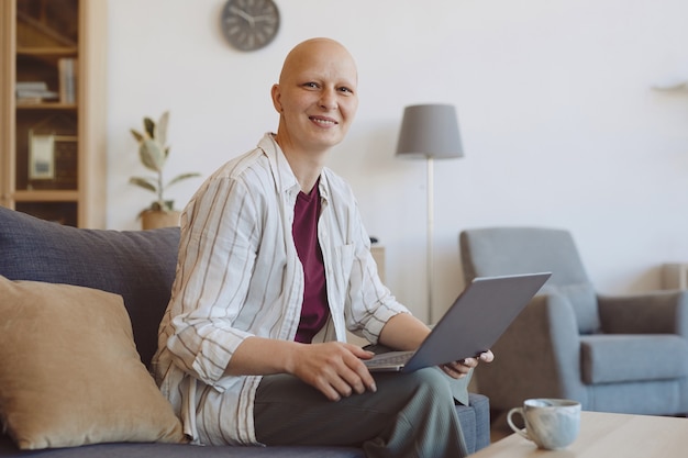 Retrato de uma mulher adulta careca sorrindo para a câmera enquanto está usando o laptop sentado no sofá no interior de uma casa moderna, alopecia e conscientização do câncer, copie o espaço