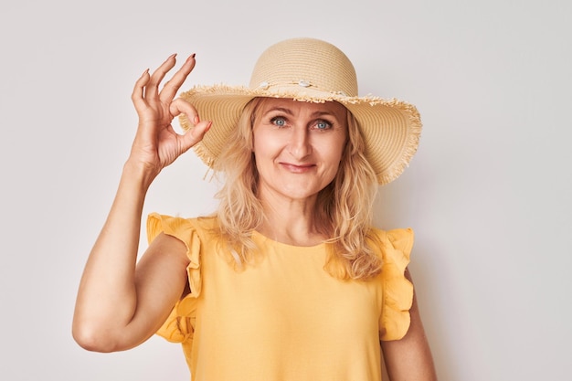 Retrato de uma mulher adulta de chapéu e vestido amarelo mostrando sinal de OK com os dedos isolados no fundo branco Conceito de férias de carreira bem-sucedida