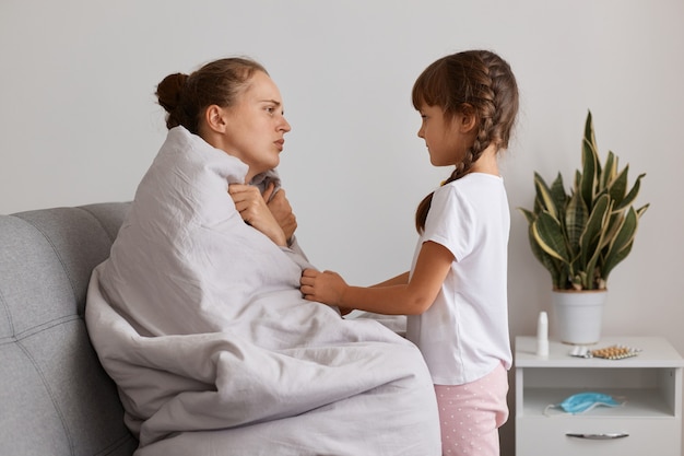 Retrato de uma mulher adulta jovem triste, sentada no sofá, embrulhada em uma manta, sentindo frio, sofrendo de gripe, linda filha de cabelos escuros com rabo de cavalo em pé perto da mãe, quer ajudar a mamãe doente.