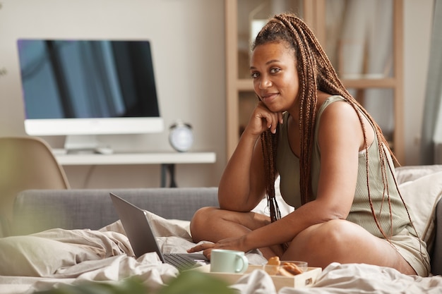 Retrato de uma mulher africana com um penteado elegante, enquanto toma o café da manhã e usa o laptop na cama dela pela manhã