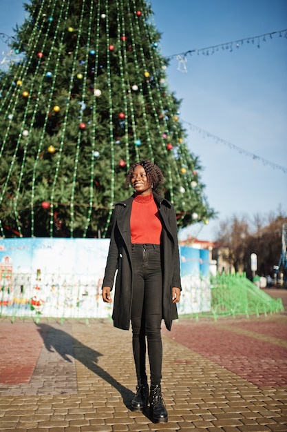 Retrato de uma mulher africana de cabelos cacheados, vestindo um casaco preto elegante e uma gola alta vermelha posando ao ar livre contra a árvore principal do ano novo da cidade.