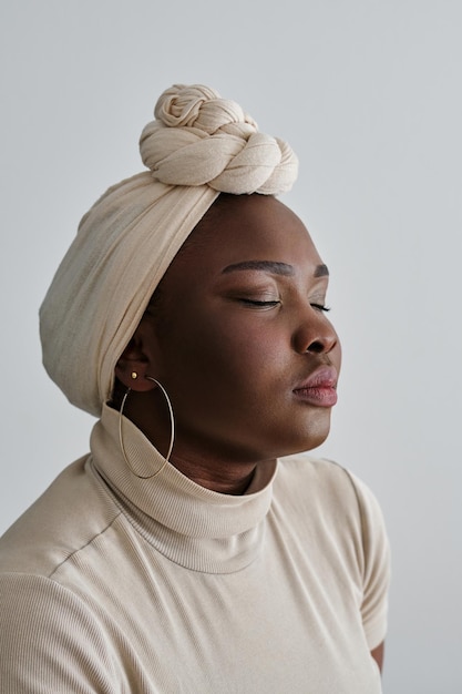 Retrato de uma mulher afro-americana calma em turbante étnico bege com os olhos fechados em fundo cinzento
